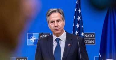 Antony Blinken in a blue suit purses his lips in front of an American flag and a NATO symbol.