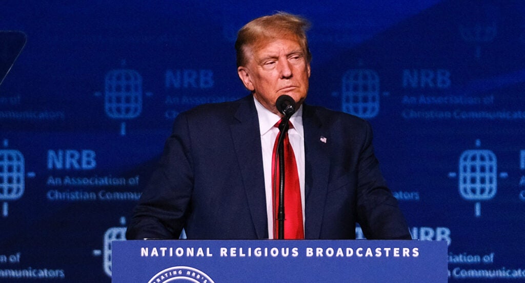 Donald Trump in a blue suit with a red tie stands behind a podium reading "National Religious Broadcasters"