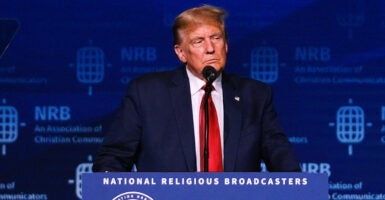 Donald Trump in a blue suit with a red tie stands behind a podium reading 