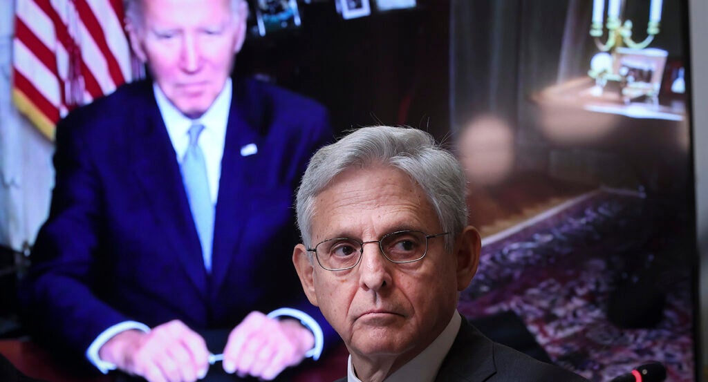 Attorney General Merrick Garland in a suit in front of a screen showing President Joe Biden in a blue suit.