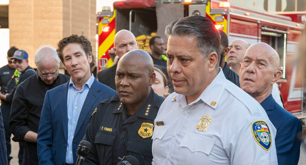 Joel Osteen in a blue suit listens as Fire Department Chief Samuel Pena and Mayor John Whitmire speak at a press conference