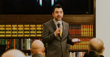 Lucas Miles in a gray suit speaks in front of bookshelves while holding a microphone