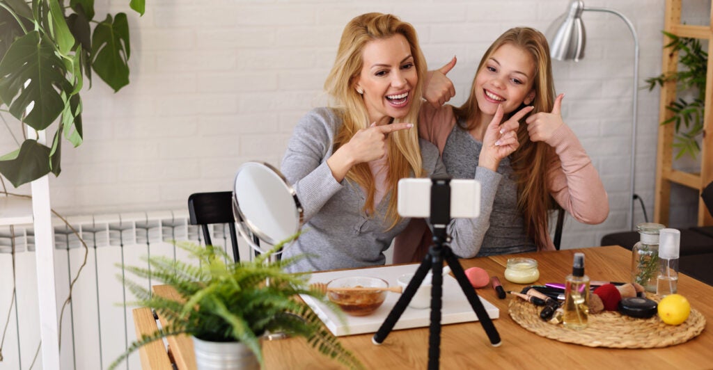 A blonde mother and daughter sit at a kitchen table, smiling and giving a thumbs up to the camera in front of them. They're both wearing grey shirts.