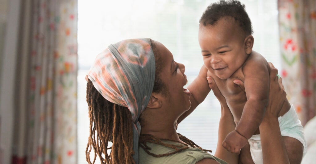 A Black woman lifts her baby up, while wearing a colorful headband. The mother has dreads, and the baby is wearing only a white diaper.