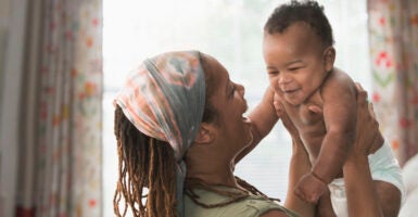 A Black woman lifts her baby up, while wearing a colorful headband. The mother has dreads, and the baby is wearing only a white diaper.