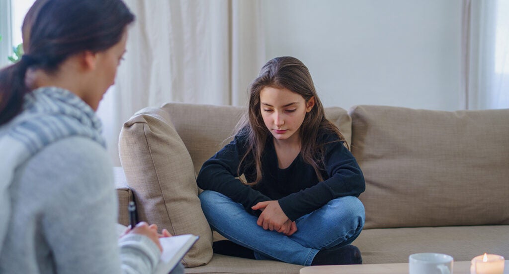 Preteen girl in jeans and a sweater crosses her hands on a couch as a therapist asks her questions with a pad and paper.