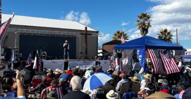 A large group of Americans sit on lawn chairs outside and listen to a speaker on state.