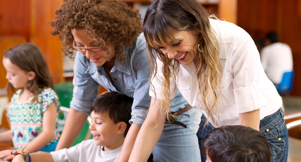 Two teachers, one wearing white and one wearing a blue jean shirt, help children learn