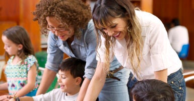 Two teachers, one wearing white and one wearing a blue jean shirt, help children learn