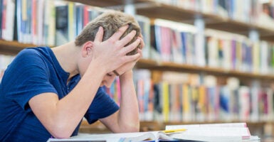 A teenage boy sits at a table, hands grabbing his hair in frustration. He is looking at a book.