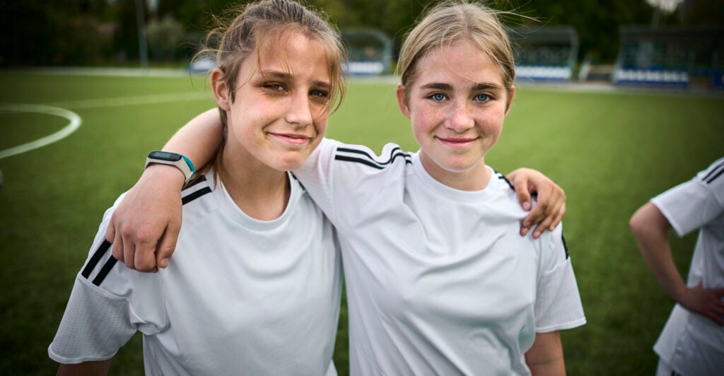 Two young girls smile for a picture, in white soccer jerseys while on a green field.