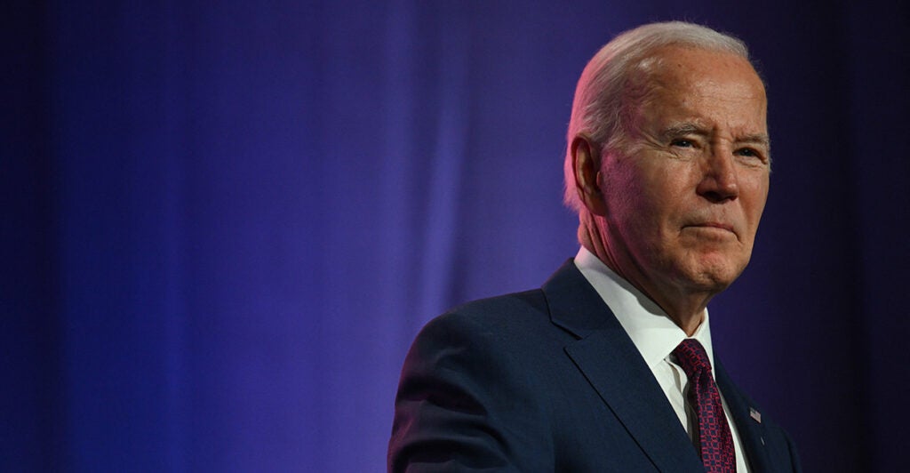 President Joe Biden wears a suit and tie on stage with a blue background behind him.