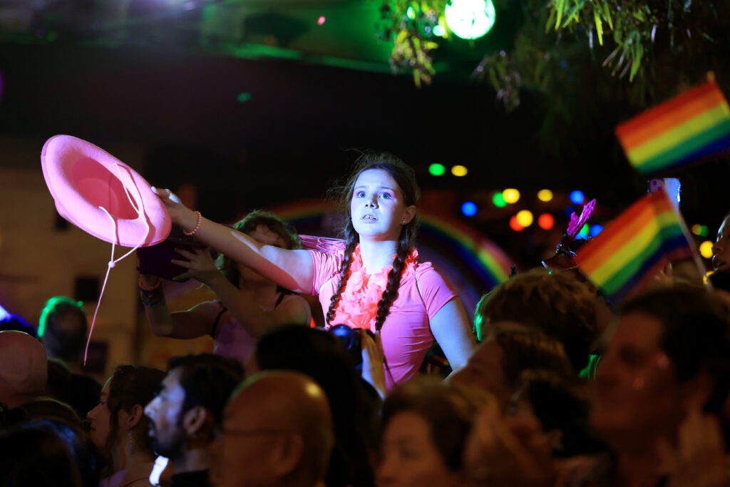 A young girl dressed in a pink shirt, holding a pink cowgirl hat, looks off into the distance at a gay pride event.