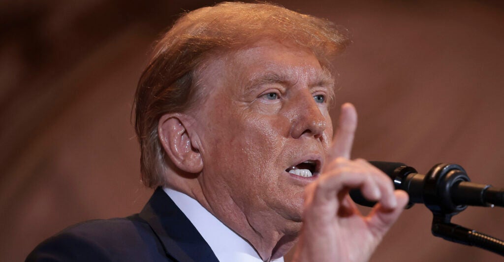 Former President Donald Trump in a suit at a microphone with his hand raised, Finger pointing upward