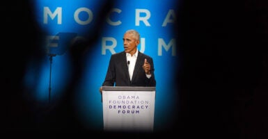 Former President Barack Obama stands at a podium and speaks in a black suit and white shirt without a tie.