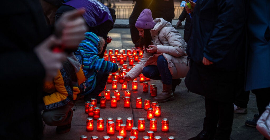 People in winter coats lights small candles in red votives on the sidewalk.