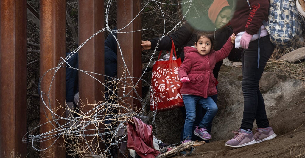 small child and women immigrants walking away from steel southern border wall and barbed wire