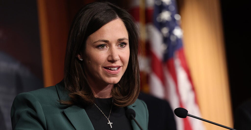 Senator Katie Britt in a dark suit at a microphone at a press conference