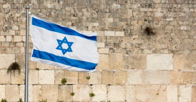 The white and blue flag of Israel flies in front of the stone Western wall.