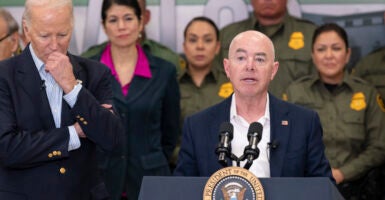 Alejandro Mayorkas stands behind a lectern in a navy blue suit, while President Joe Biden stands to the left of him.