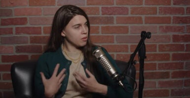 Detransitioner Camille Kiefel with her brown hair sits in front of a black microphone, while wearing a tan shirt and green sweater.