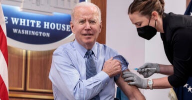 President Joe Biden receives a COVID-19 vaccine shot from a nurse while sitting down with his suit jacket off.