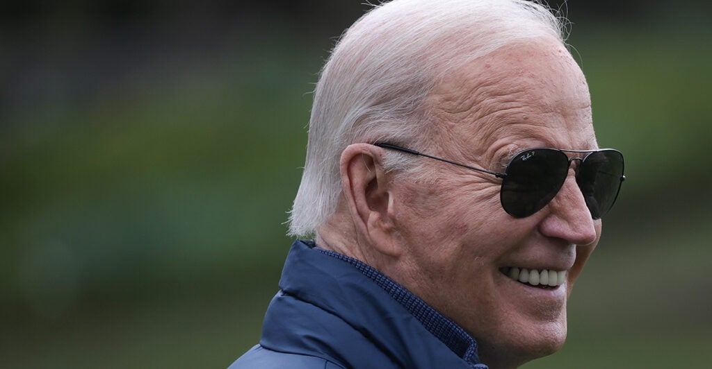 President Joe Biden smiles in a navy jacket or vest and Aviator sunglasses.