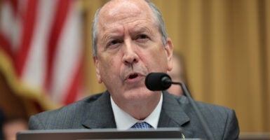 Rep. Dan Bishop speaks into a microphone while wearing a grey suit. An American flag is seen behind him.