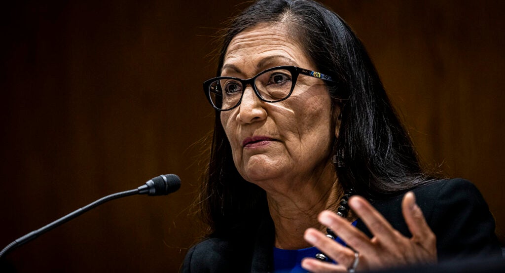 Interior Secretary Deb Haaland looks on, gesturing in front of a microphone and wearing a blue dress with a black suit jacket and pearls.