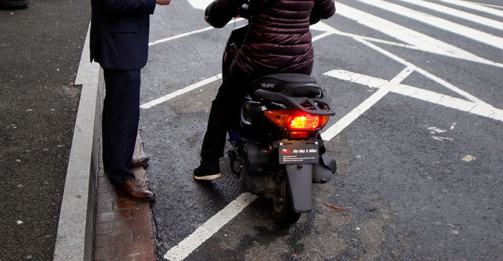 A man stands on the street talking to a woman on a motor-driven cycle.