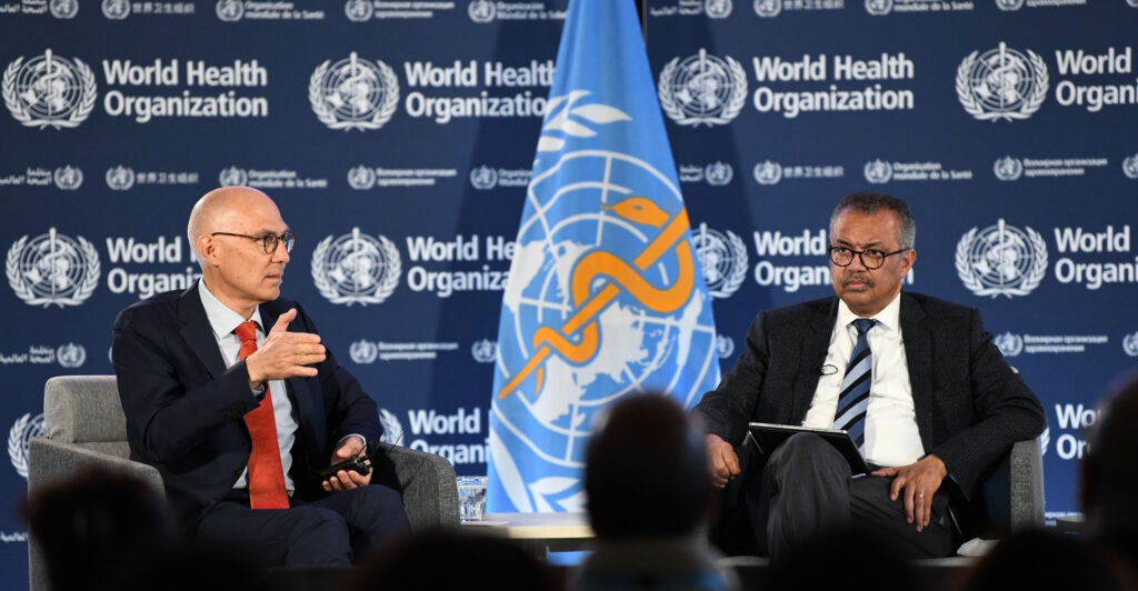 Two elderly men sit in chairs while wearing black suits as a blue flag stands in between them.