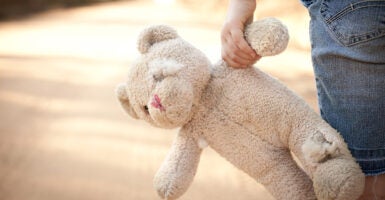 A child walks down the street holding a dirty white teddy bear.