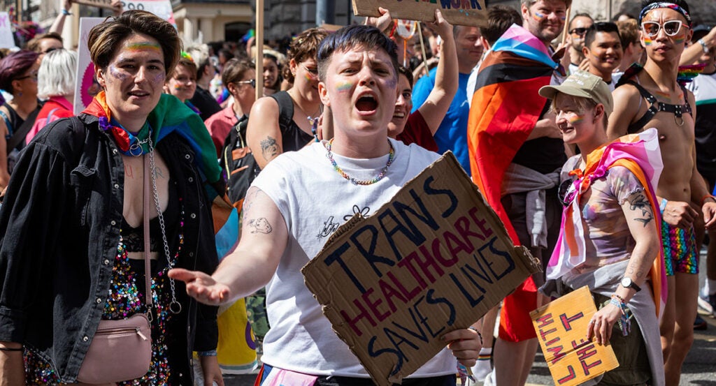 Protesters march, carying a sign reading, "Trans Health Care Saves Lives"