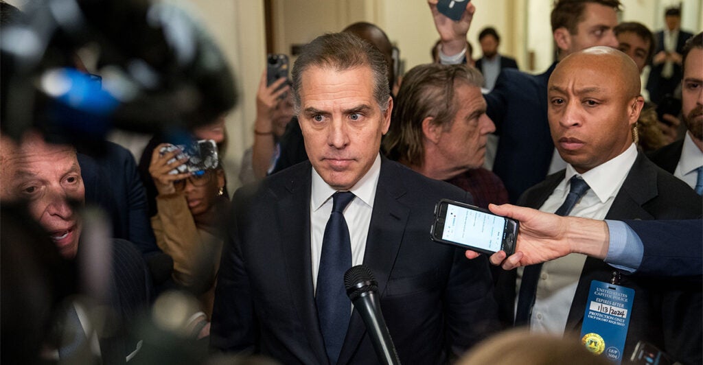 Hunter Biden stands in the middle of a crowd with microphones and cameras with a stern look on his face.