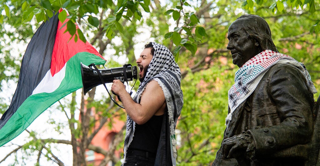 Male pro-Palestinian protester with Palestinian flag, headdress, and a megaphone alongside a bronze statue of Benjamin Franklin