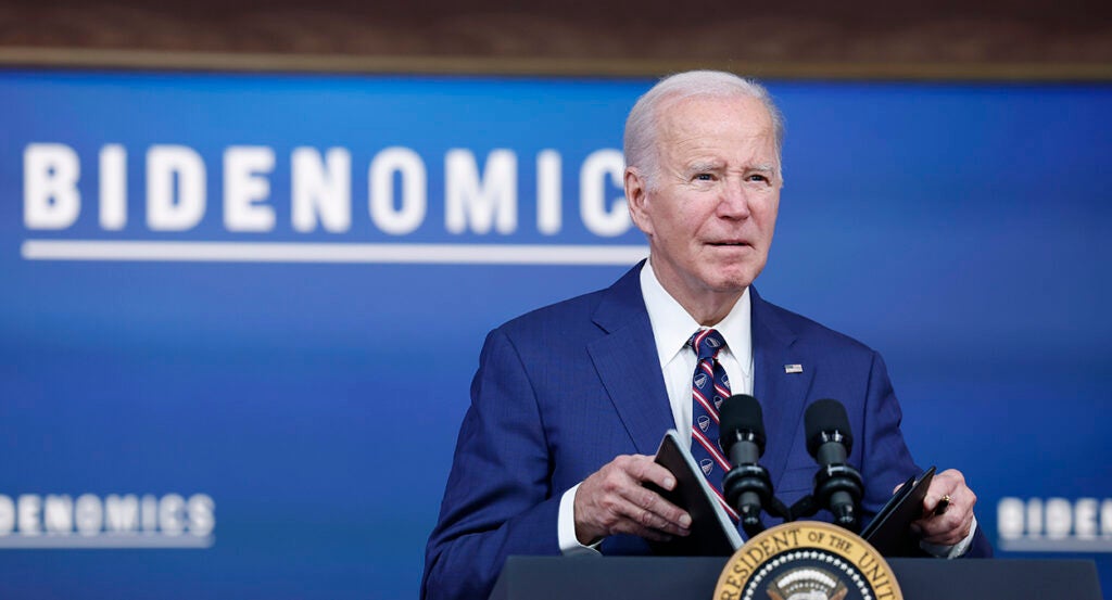 Joe Biden in a blue suit holds a briefing book behind the presidential seal and in front of a sign reading "Bidenomics."