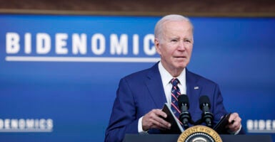 Joe Biden in a blue suit holds a briefing book behind the presidential seal and in front of a sign reading "Bidenomics."