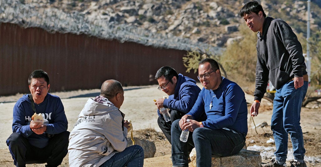 Chinese immigrants sit on the ground next to the U.S. border wall