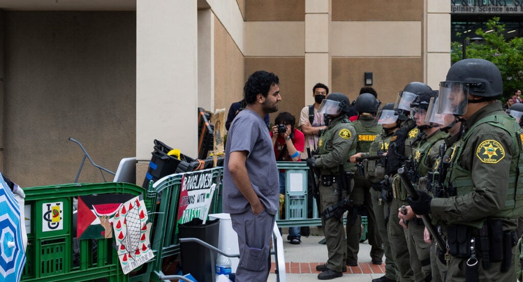 A protester wearing a gray shirt and jeans stands before a line of police officers wearing face shields and green uniforms.