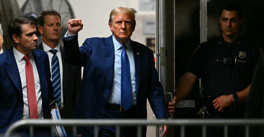 Donald Trump in the suit with his fist in the air surrounded by lawyers and court police