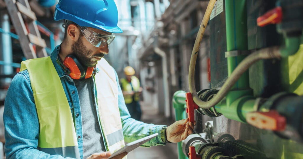 A young male engineer With a hard hat is examining the pipe system In a factory and checking the data on his tablet.