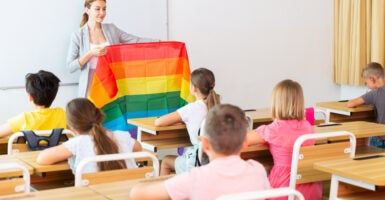 Young progressive female teacher discussing with preteen children about LGBT social movements in classroom, holding rainbow flag.