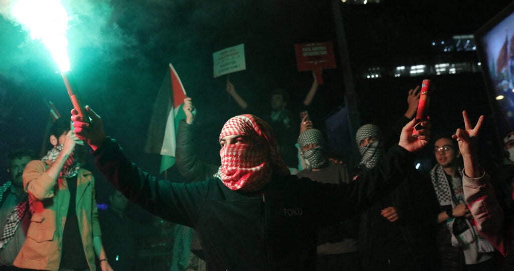 A man wearing a head and face covering holds a green flare in the air.