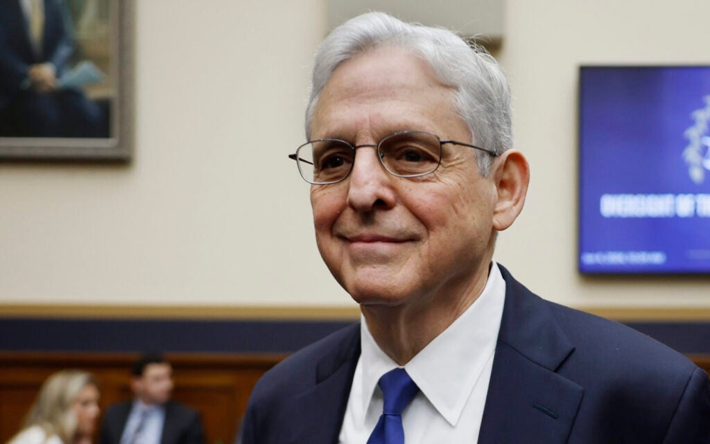 Attorney General Merrick Garland smiles as he enters a room, wearing a blue suit and tie.