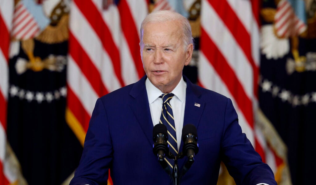 Joe Biden wears a navy-blue suit and speaks at a podium in front of American flags.