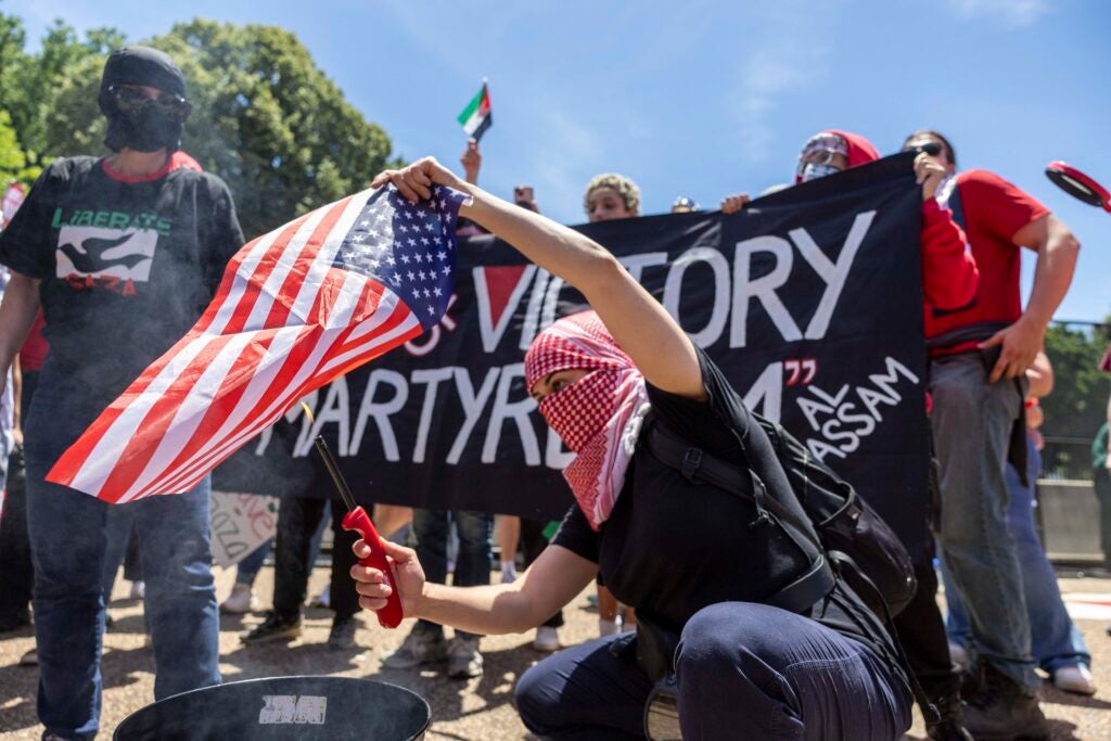 A man wearing a head and face covering crouches as he lights an American flag on fire over a bucket.