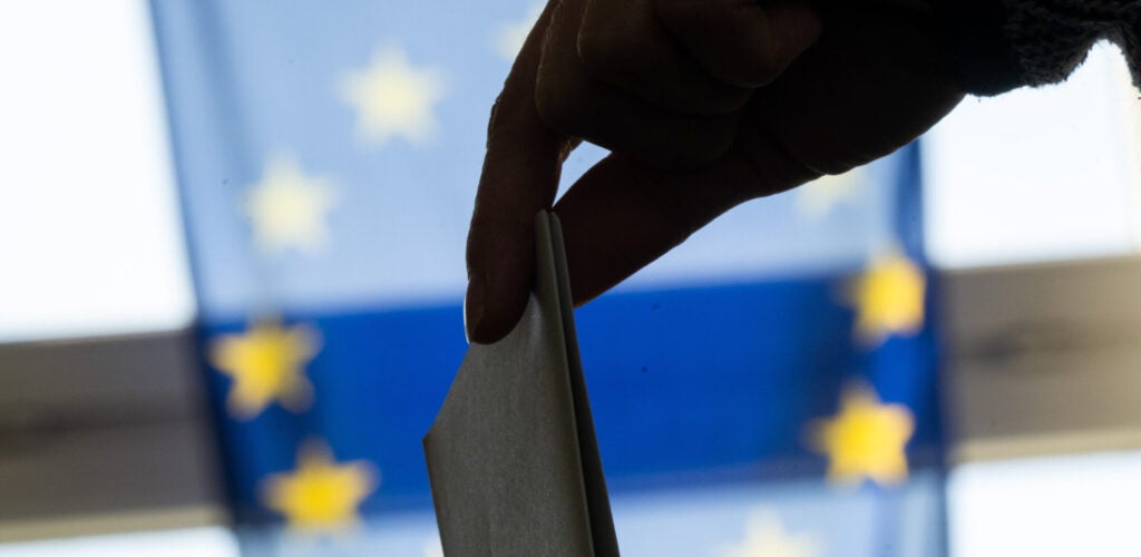 A woman's hand drops a paper ballot into a ballot box in front of the European Union flag.