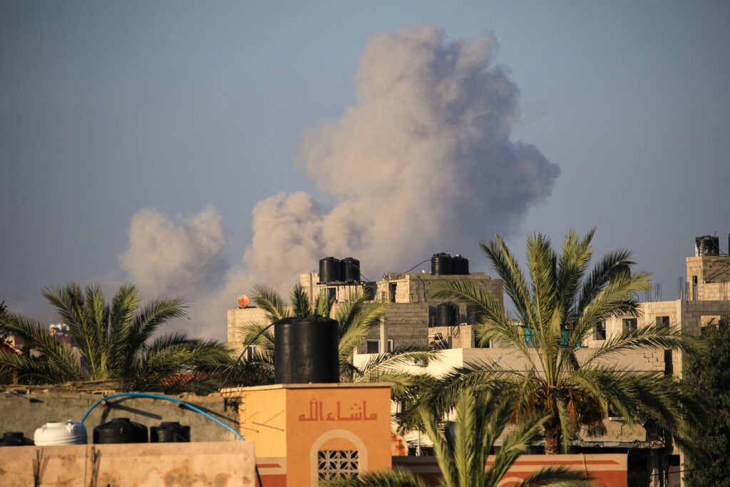 Smoke billows overhead the tops of buildings and palm trees.