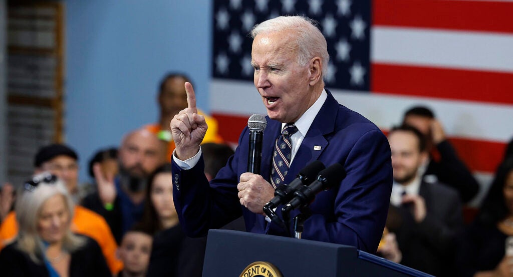 President Joe Biden in a blue suit raises a finger while speaking in a microphone