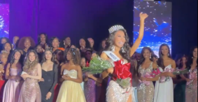 Trans-identifying male contestant waves waves from the stage in a gown, crown, and flowers with other contestants in the background